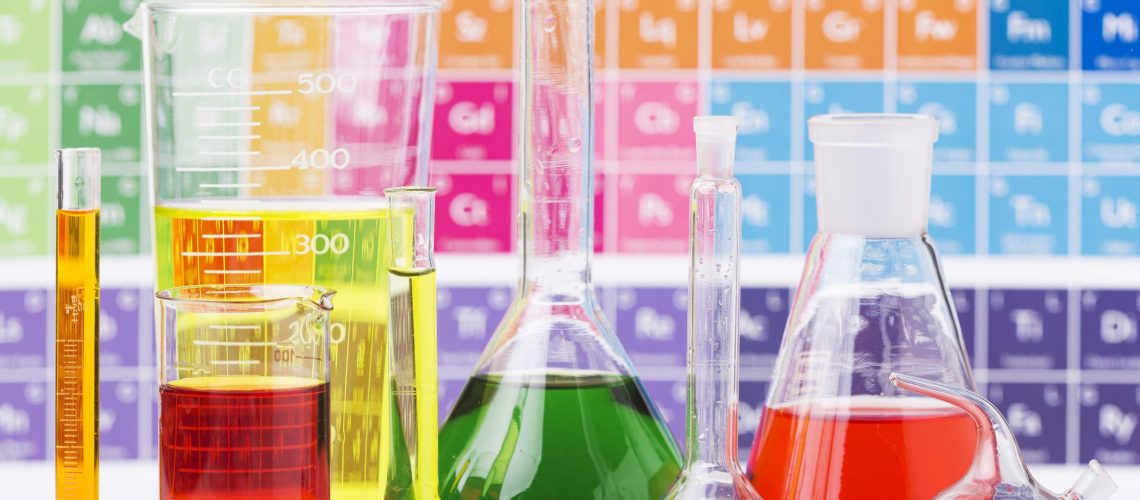 inorganic chemicals in flasks and beakers in front of a period table of elements