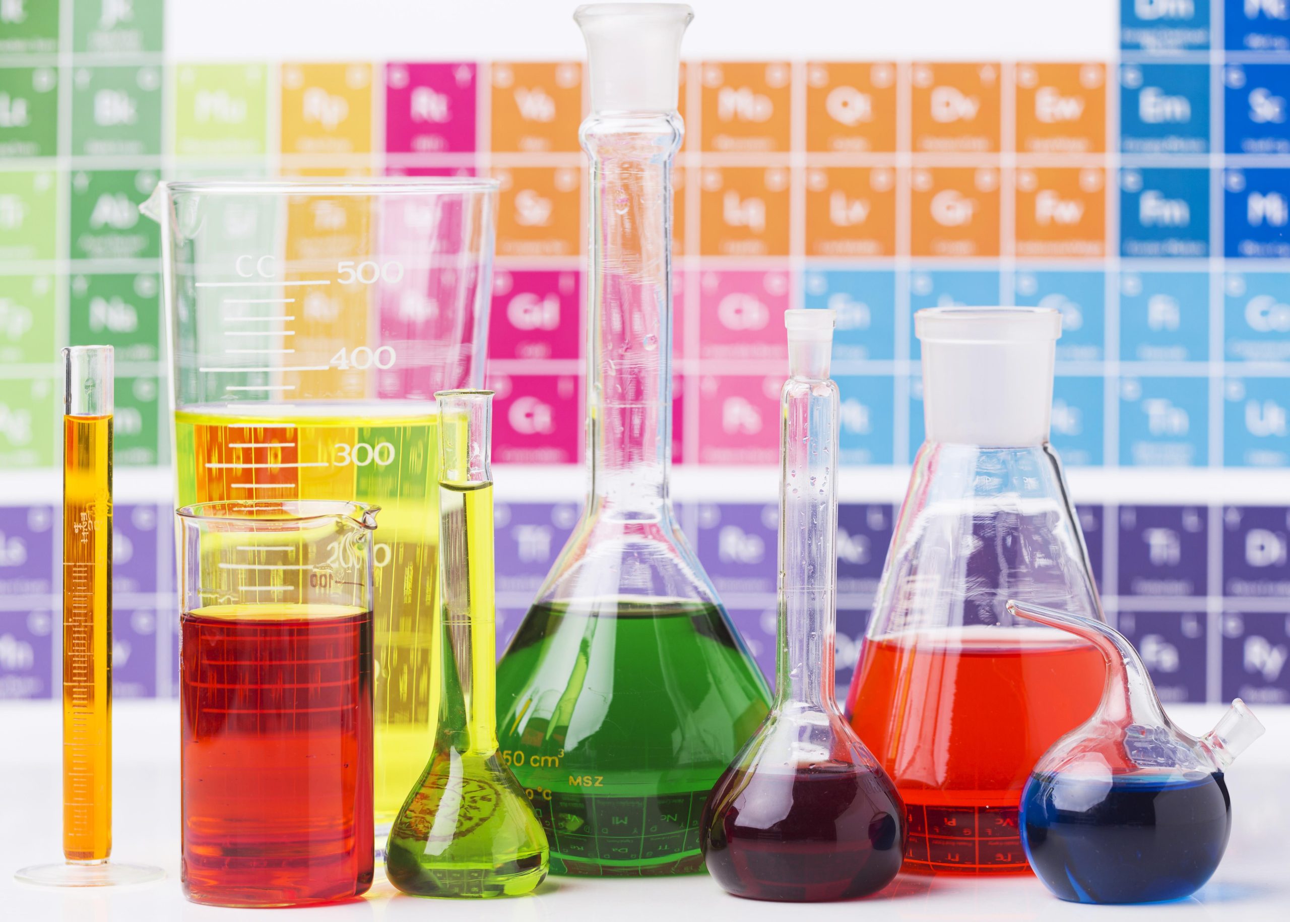 inorganic chemicals in flasks and beakers in front of a period table of elements